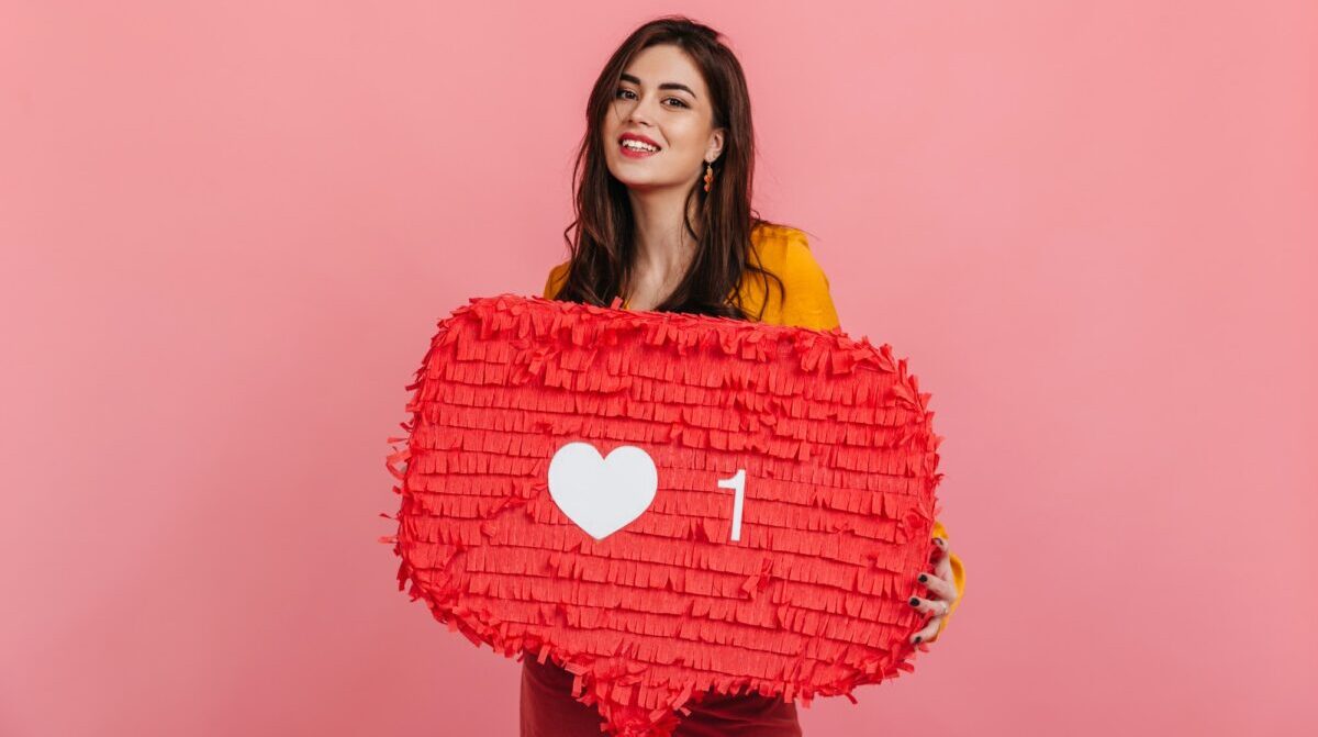 Teenager girl in bright outfit is smiling, holding red "Like" sign from Instagram on pink backgroun